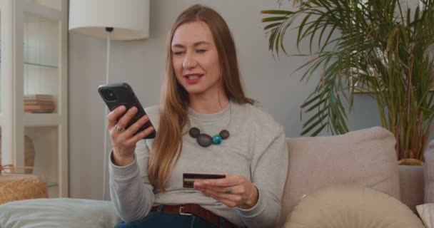 Feliz animado atraente jovem empresária lê e insere detalhes do cartão de crédito no aplicativo de pagamento de smartphones em casa. — Vídeo de Stock