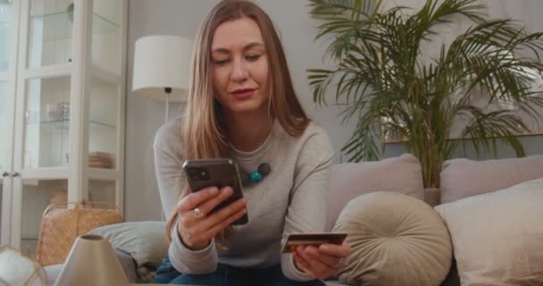 Piękna szczęśliwa blondynka biznes kobieta czyta, pisze w plastikowych szczegółach karty w smartfonie zakupy aplikacji w lekkim domu. — Wideo stockowe