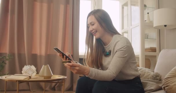 Happy smiling young woman reads card to pay online via smartphone app, enjoys shopping online at light spacy apartment. — Stock Video