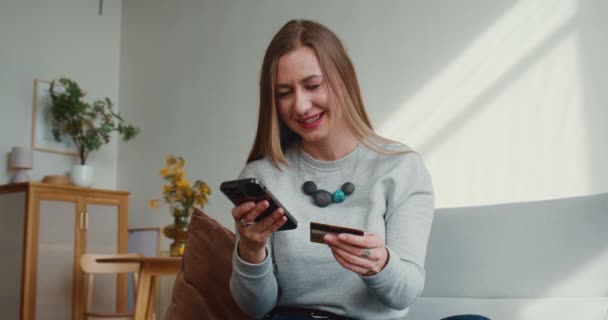Όμορφη ευτυχισμένη νεαρή γυναίκα εισάγει τον αριθμό της κάρτας στην εφαρμογή πληρωμής για ψώνια smartphone, γέρνει πίσω χαρούμενα στο σπίτι φως. — Αρχείο Βίντεο