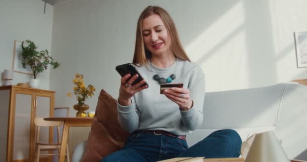 Online koncepció vásárlása. Gyönyörű boldog fiatal mosolygós nő belép hitelkártya számok segítségével smartphone app otthon. — Stock videók