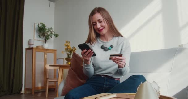 Hermosa mujer alegre feliz joven entra en los números de tarjeta de crédito utilizando la aplicación de teléfono inteligente en casa, sonrisas en la emoción. — Vídeos de Stock
