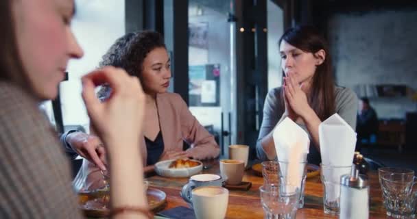 Група молодих красивих щасливих багатоетнічних жінок випадково розмовляють за модним легким столом кафе з їжею та кавою за обідом . — стокове відео