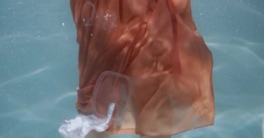 Plastik atık sorunu. Kadın bacakları suyun altında duruyor, çeşitli tek kullanımlık ambalaj çöpleri yavaş çekim etrafında yüzüyor..