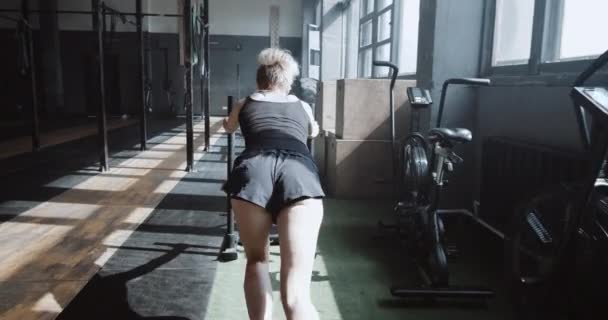 Pokonywanie trudności. Powrót widok młoda atletyczna kobieta pchanie treningu sanki ćwiczenia w dużej pustej siłowni slow motion. — Wideo stockowe
