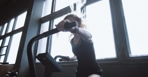 Giovane bella donna bionda atletica che lavora su una macchina da ginnastica fissa con vento nei capelli dal fan slow motion. — Video Stock
