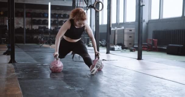 Giovane professionista atletica dai capelli rossi allenatore donna facendo split stretch esercizio su kettlebells in grande palestra rallentatore. — Video Stock