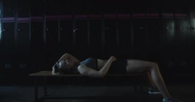 Yorgun atletik sarışın kadın karanlık spor salonunun soyunma odasında bankta uzanmış, ayağa kalkıyor ve ağır çekimde devam ediyor..