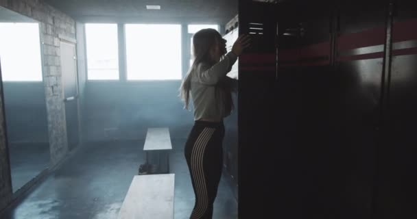 Camera follows sad athletic blonde woman entering dark gym locker room angry, tired and upset after bad day slow motion. — Stock Video