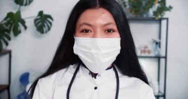 Yakın plan mutlu, melez, yüz maskesi takan kadın doktor, laboratuvar önlüğü, açık modern klinik ofisinde kameraya gülümsüyor.