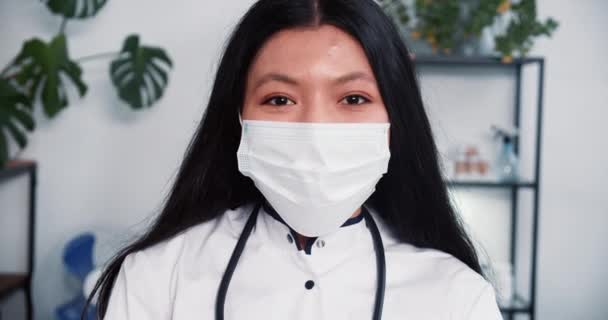 Close-up jovem feliz misto raça fêmea médico vestindo máscara facial, jaleco sorri para a câmera no escritório clínica moderna luz — Vídeo de Stock