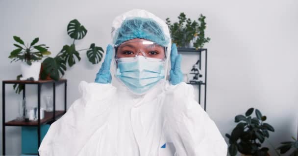 Jovem médica multiétnica usa terno médico descartável e máscara, ajustando óculos durante o surto de pandemia. — Vídeo de Stock