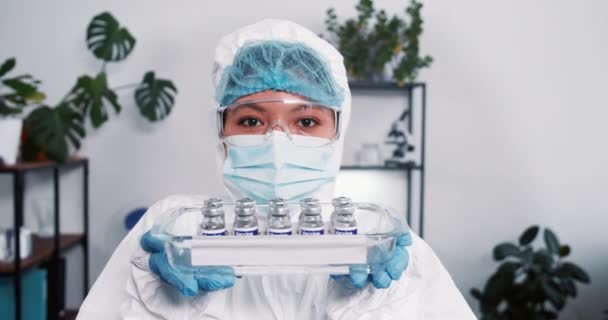 Победа над коронавирусом. Веселая женщина-лаборантка в защитном костюме показывает медицинский поднос с фляжками от вакцины. — стоковое видео