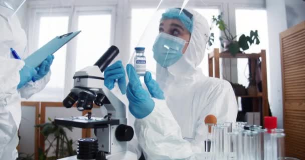Próby szczepionek. Profesjonalny lekarz apteki, naukowiec w pełnym kombinezonie ochronnym posiada kolbę z lekiem w laboratorium. — Wideo stockowe