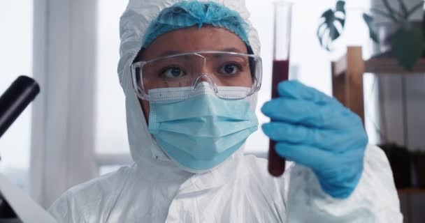 Ensaios clínicos. Jovem focada raça mista cientista médico de laboratório farmacêutico em terno de proteção olhando para tubo de teste com sangue. — Vídeo de Stock