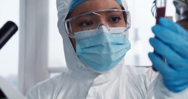 Лаборант за работой. Молодой серьезный многонациональный вирусолог врач женщина в защитном костюме проведение пробирки с кровью — стоковое видео
