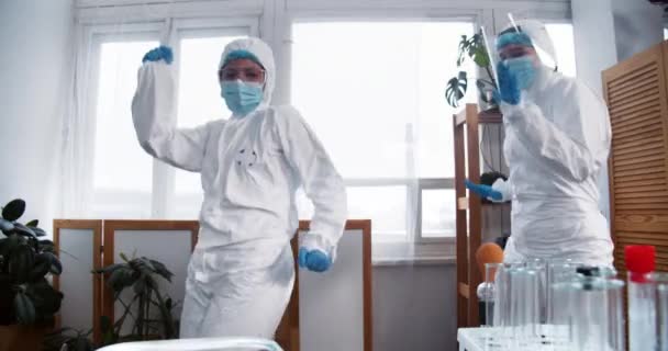 Dois cientistas divertidos felizes do sexo feminino, médicos em ternos de proteção médica branca dança comemorando o sucesso da pesquisa no laboratório. — Vídeo de Stock