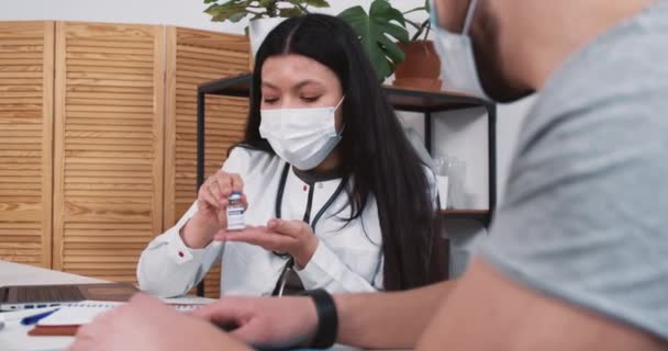 Fröhliche Mischlingshündin Apothekenärztin im Laborkittel zeigt männlichem Patienten Impfflasche am lichten Kliniktisch. — Stockvideo