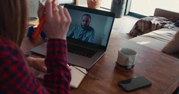 Webcam videogesprek. Achteraanzicht jonge blonde vrouw praten met gelukkig mannelijke vriend met behulp van laptop, het maken van notities aan huis tafel. — Stockvideo