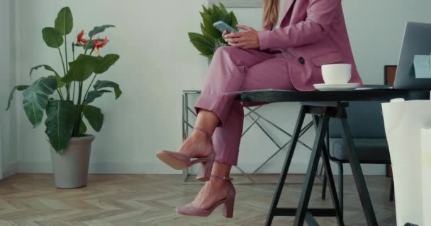 Närbild elegant affärskvinna i rosa kostym med hjälp av smart telefon app med kaffe sittbenen korsade på kontorsbordet. — Stockvideo