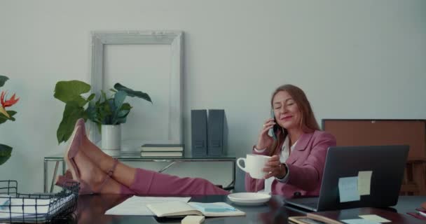 Belle heureux succès patron féminin sourit parler sur téléphone mobile, sirotant du café avec les jambes sur la table de travail de bureau. — Video