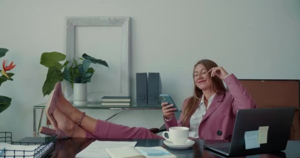 행복하고 아름다운 여성 사장 이 사무실 테이블에 다리를 놓고 웃으며 휴대폰 앱을 이용해 온라인으로 물건을 사는 모습. — 비디오