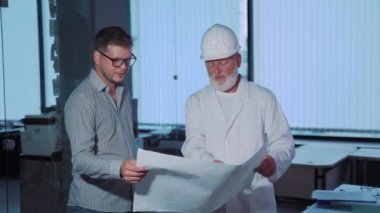 Deneyimli bir erkek inşaat mühendisi bir meslektaşıyla yaptığı toplantıda çizimlere bakar.