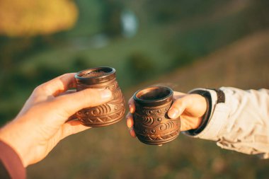 Sabahları iki bardak sıcak içeceği doğada tutan iki elin resmi.