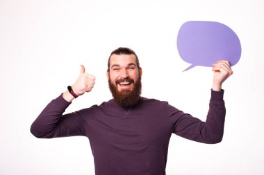 Genç sakallı bir erkek kameraya gülümsüyor ve elinde bir konuşma balonu tutuyor.