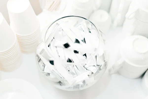 Una foto de paquetes de azúcar blanco en una canasta cerca de unas tazas blancas — Foto de Stock