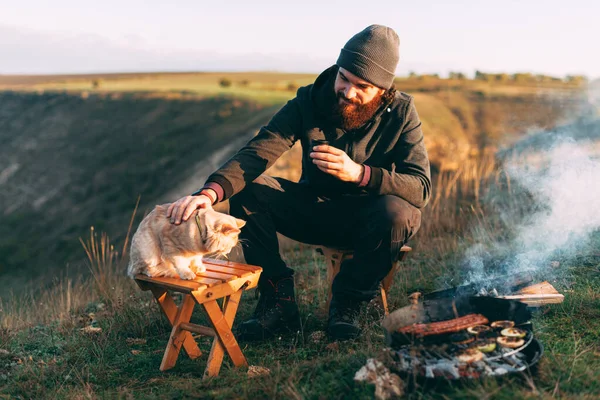 Μια φωτογραφία ενός νεαρού άνδρα που κάθεται σε ένα λόφο με τη γάτα του κοντά σε μια ψησταριά με έναν καφέ στο χέρι του — Φωτογραφία Αρχείου