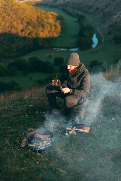 Γενειοφόρος νεαρός άνδρας σε ένα λόφο κάθεται κοντά σε μια σχάρα με έναν καφέ — Φωτογραφία Αρχείου