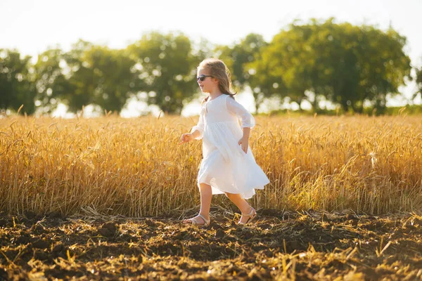 Fröhliches kleines Mädchen mit weißem Kleid und Sonnenbrille läuft lachend über ein Weizenfeld — Stockfoto