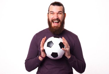 Genç sakallı adam kameraya heyecanlı bakıyor ve elinde bir futbol topu tutuyor.