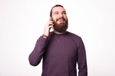 Sakallı genç bir adam gülümsüyor, başka tarafa bakıyor ve telefonuyla konuşuyor.