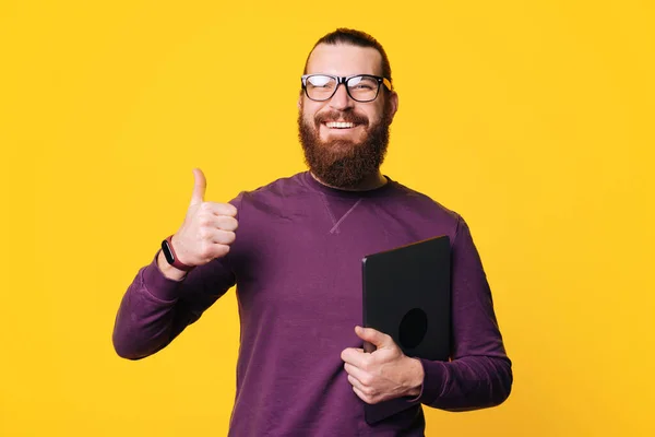 Un jeune homme barbu joyeux tient un ordinateur montrant un pouce levé porte des lunettes près d'un mur jaune — Photo