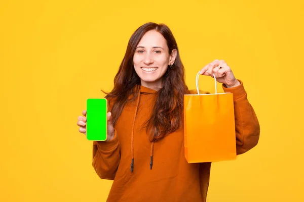 Egy vidám fiatal nő egy bevásárló táskát és egy telefont tart a kezében. — Stock Fotó