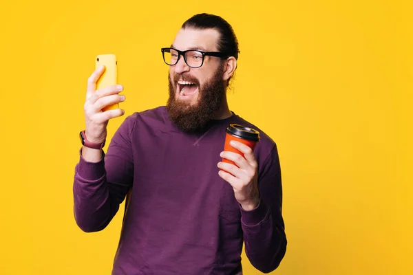 Бородатий молодий чоловік в окулярах дивиться збуджено на свій телефон і тримає чашку з гарячим напоєм — стокове фото