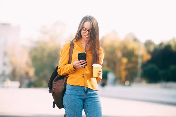 Una foto de una mujer con una taza de bebida caliente en la mano y sosteniendo un teléfono está caminando por una ciudad — Foto de Stock