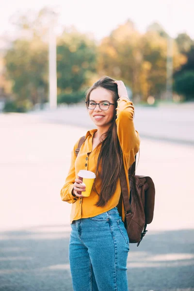 Retrato de uma jovem mulher usando óculos no parque e segurando uma xícara de bebida quente sorrindo para a câmera — Fotografia de Stock