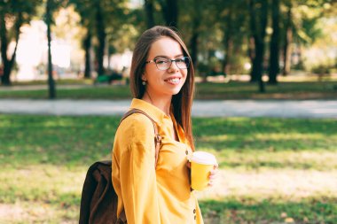 Gözlüklü bir kadın parkta elinde bir bardak sıcak içecekle kameraya gülümsüyor.