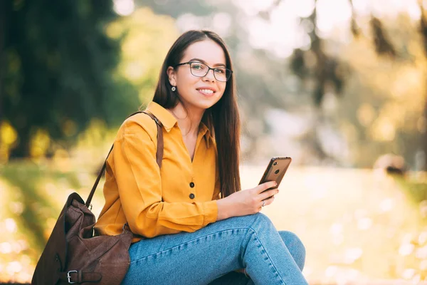 Uma jovem alegre olhando e sorrindo para a câmera está segurando um telefone no parque — Fotografia de Stock