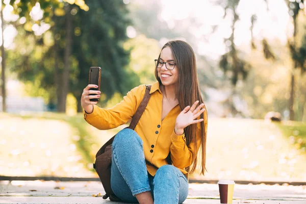 Una joven con gafas sonríe y agita su teléfono sentado en algún lugar de la naturaleza afuera. — Foto de Stock