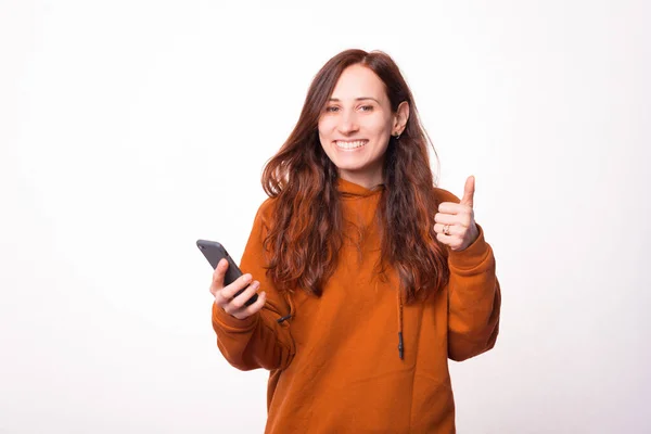 Boldog fiatal nő kezében egy telefon és mutatja a hüvelykujját felfelé mosolyog a kamerába — Stock Fotó