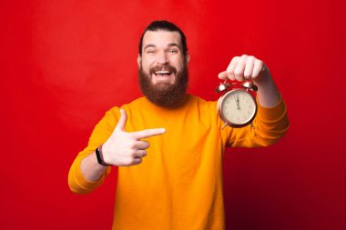 Genç sakallı adam kameraya mutlu bir şekilde bakıyor ve elinde bir saat tutuyor.