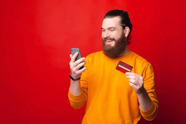 Feliz joven barbudo mirando en su teléfono y sosteniendo una tarjeta de crédito — Foto de Stock