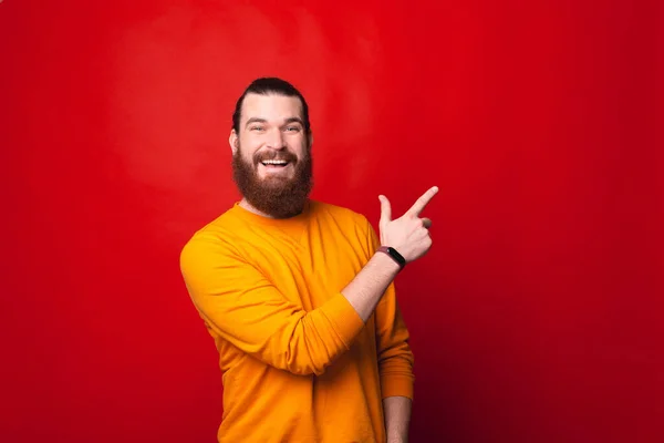 수염을 기른 한 남자가 카메라 앞에서 웃으며 붉은 벽 근처의 자유 로운 공간을 가리키고 있다 — 스톡 사진