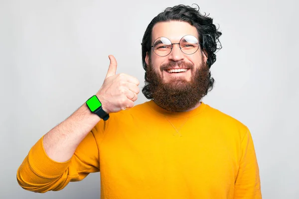 Gelukkig bebaarde hipster man met duim omhoog en het dragen van slimme horloge met leeg groen scherm — Stockfoto