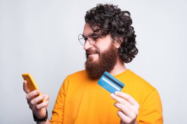 Sakallı neşeli genç adam elinde telefonu ve kredi kartıyla internetten alışveriş yapıyor.