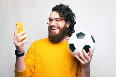 Genç sakallı bir adam telefonuna gülümsüyor ve elinde bir futbol topu tutuyor.